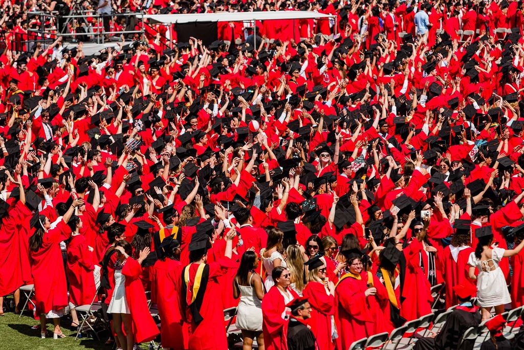 Shot of students on Nickerson field during commencement. They are wear red gowns and black mortarboards and raise their hands up in celebration during the ceremony.