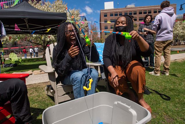 Photo of Jordana Kasaato (ENG’23), left, and Kendall Lowe (CAS’22) try their hand at a fishing game on the COM lawn during the BU Brazilian Association’s Junina Party in honor of the annual Festas Juninas April 30, which in Brazil runs throughout June. Kasaato, a young Black woman with long braids, who wears a black jacket and jeans, laughs as her fishing line gets tangled with Lowe's. Lowe is a young Black woman with long braised who wears a black sweater and amber pants. She smiles as she holds up her fishing rod. The pair are sitting on lawn chairs and fishing from a large plastic bin filled with water. Other students on the COM lawn can be seen behind them, it's a sunny spring day.