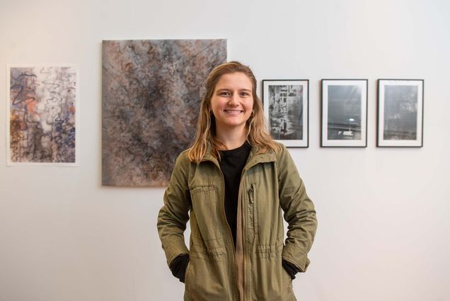 Photo of Brianna Howard (CFA’22) standing in front of her work at the BFA Thesis Art Exhibit at Stone Gallery May 5. Brianna is white woman with blonde hair; she smiles with her hands in her pockets, wears a green jacket and black plants. From left, they are named Reposition, Overlapped, Proximity, Talking with the Shadows, and Remnant. The prints are mostly gray and tan colors and are abstract in nature, featuring long curving lines.
