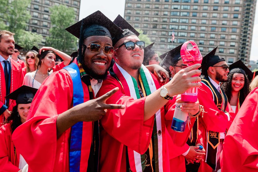 Photo of two students using a pink mister to cool off during commencement. At right, a young Black man smiles, wears sunglasses, and makes the peace sign with his right hand. At right, a young light-skinned man who holds the mister and smiles. They both wear caps and gowns.