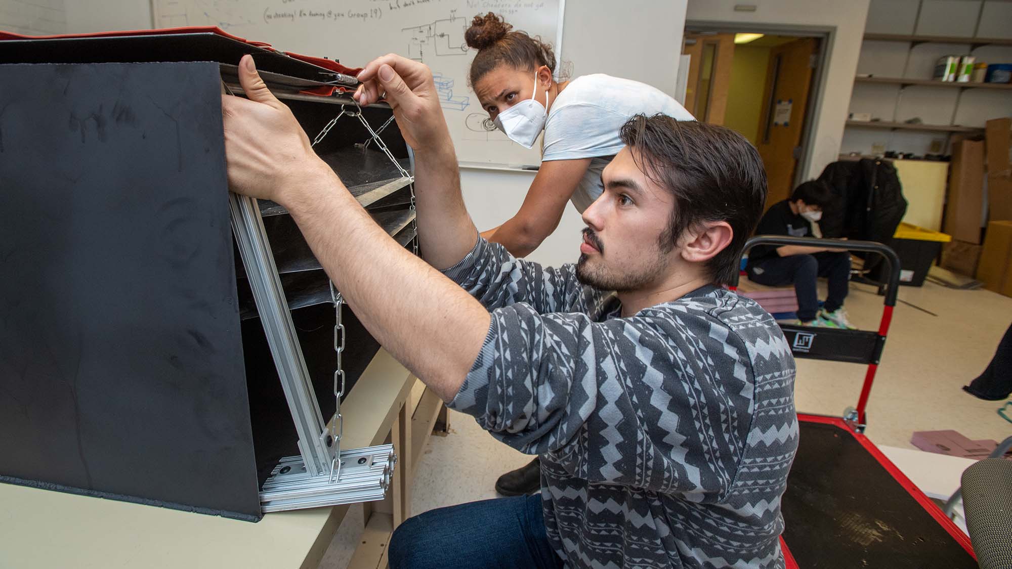 Photo of Tess Ravick (ENG’22), back, and Trevor Melsheimer (ENG’22), front work on their senior design project April 29. The device harvests water from the air. Ravick has light brown skin, wears a t-shirt, a white face mask, and their hair in a bun. Melsheimer, who has light skin and a beard and wears a gray sweater, adjusts a level of the black device on the table in front of them.
