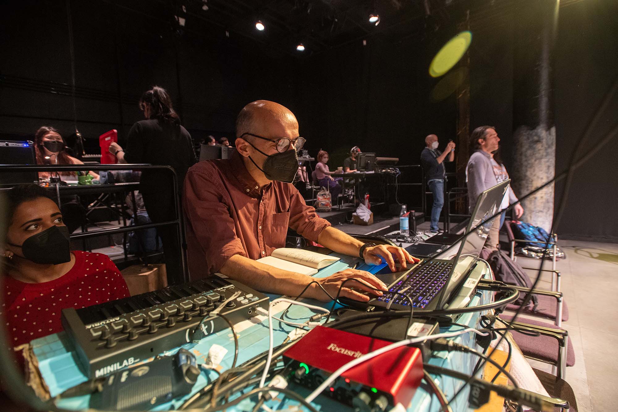 Photo of artist in residence Paolo Scoppola, right, using a laptop and checking on his software during a tech run of Exit the King April 27 at CFA’s Studio One. He stands at a table with other tech equipment, lots of wires and pre-amps are seen. Behind him, others, masked, look towards the stage.