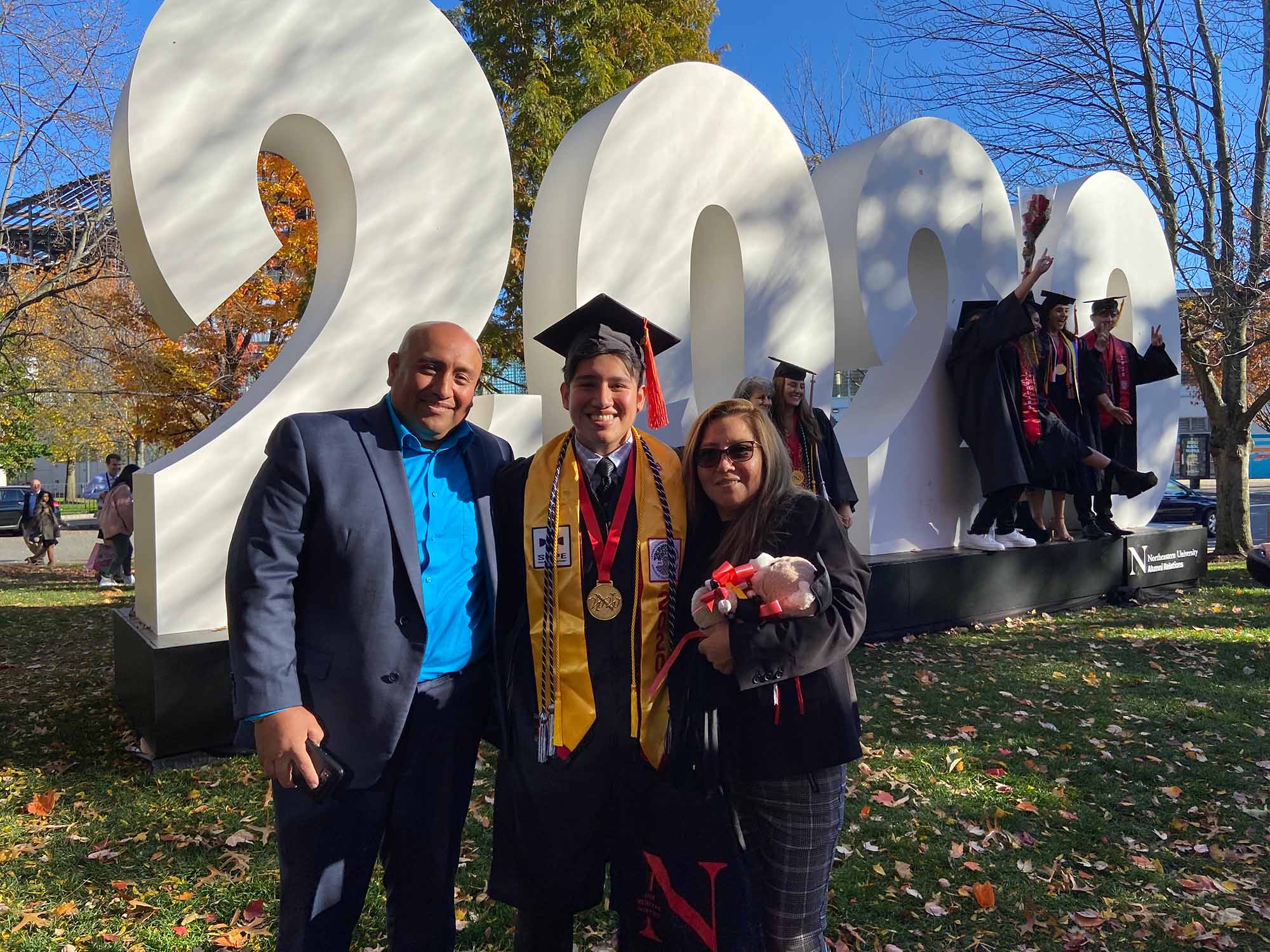 Photo of Hector Grande with his parents, Mario and Nerida, at his graduation from Northeastern University in 2020. Hector stand in a black graduation cap and gown with numerous tassels and awards around his neck/shoulders as his parents stand on either side of him.