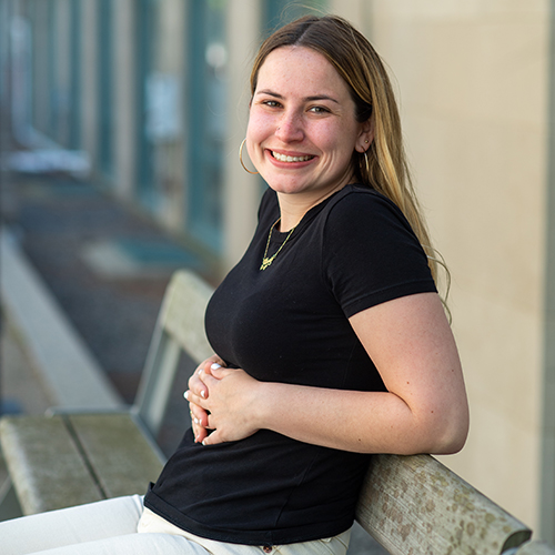 Photo of Newbury center grad Annie Rose Hallett. Hallett wears a black shirt and light tan pants. She sits on a wooden bench with hands intertwined and resting on her stomach as she turns and smiles tot he camera.