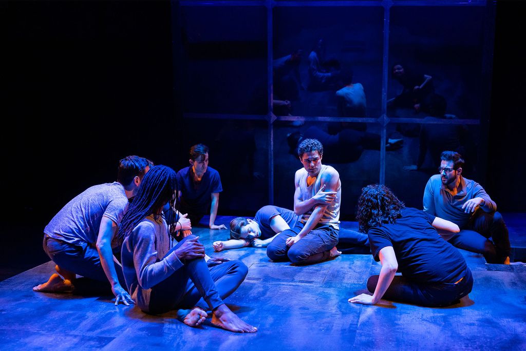 Mishka Yarovoy (CFA’23) (center) and the cast of SpeakEasy Stage’s The Inheritance. A blue light shrouds a large group of people sitting on a stage