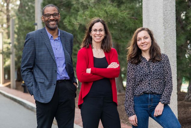 Photo of Pamela Templer (center), Shawn Jones (left), and Yasmin Romitti. From left, a tall Black man with glasses, a dark grey blazer, purpled collared shirt and black pants stands next to a shorter white woman with arms crossed wearing black blouse and pants and red cardigan, and on right a younger, shorter white woman wearing a black and white polka dot shirt and jeans. They all stand, pose, and smile next to each other.