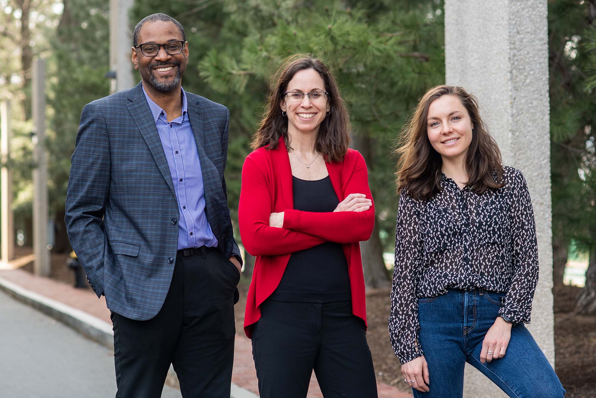 Photo of Pamela Templer (center), Shawn Jones (left), and Yasmin Romitti. From left, a tall Black man with glasses, a dark grey blazer, purpled collared shirt and black pants stands next to a shorter white woman with arms crossed wearing black blouse and pants and red cardigan, and on right a younger, shorter white woman wearing a black and white polka dot shirt and jeans. They all stand, pose, and smile next to each other.