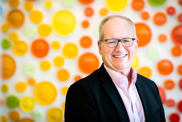 Photo of David Waters, an older white man wearing black glasses, black blazer and pants, and a pastel pink collared shirt. He is turned towards the camera and smiling as he sits in front of a blurry background featuring bright red, yellow, and orange dots on a white background.