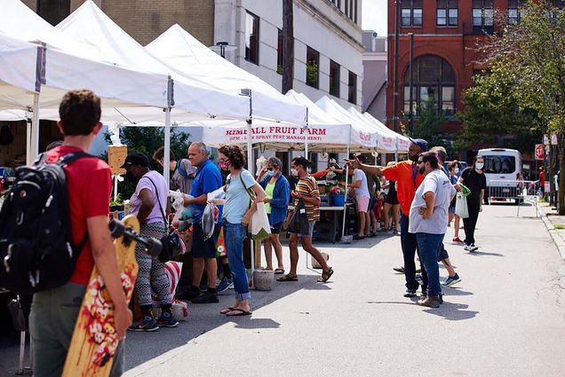 Photo of people looking at and wandering through a farmer's market on a paved road. White tent tops are set up with vendors beneath as people shop and wander on a sunny day.