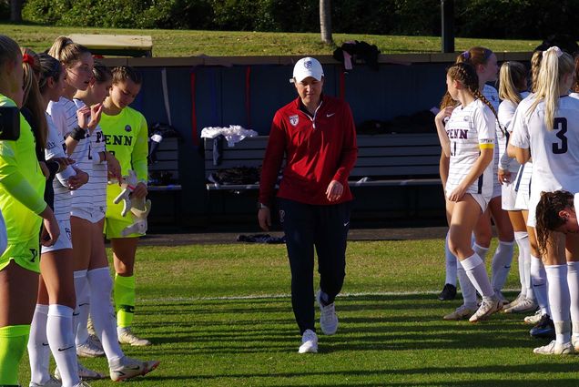 Photo of Casey Brown walking down a soccer field between two rows of Penn players in white. She wears a white cap, red pants with a Penn logo, and black pants.