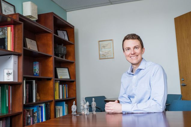 John M. Marston sits and smiles at the camera in a blue collared shirt. There is a wooden desk and bookcase nearby.