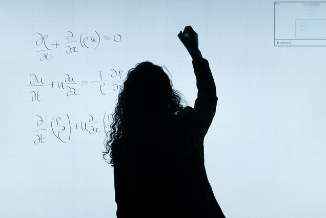 Photo of a silhouette of a person with long curly hair writing equations on a clear board