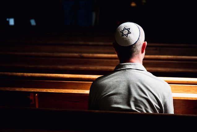 Photo taken from behind a Jewish worshipper as they sit in a synagogue. They wear a blue and white star of David kippah. The space is darkened but they are spotlighted.