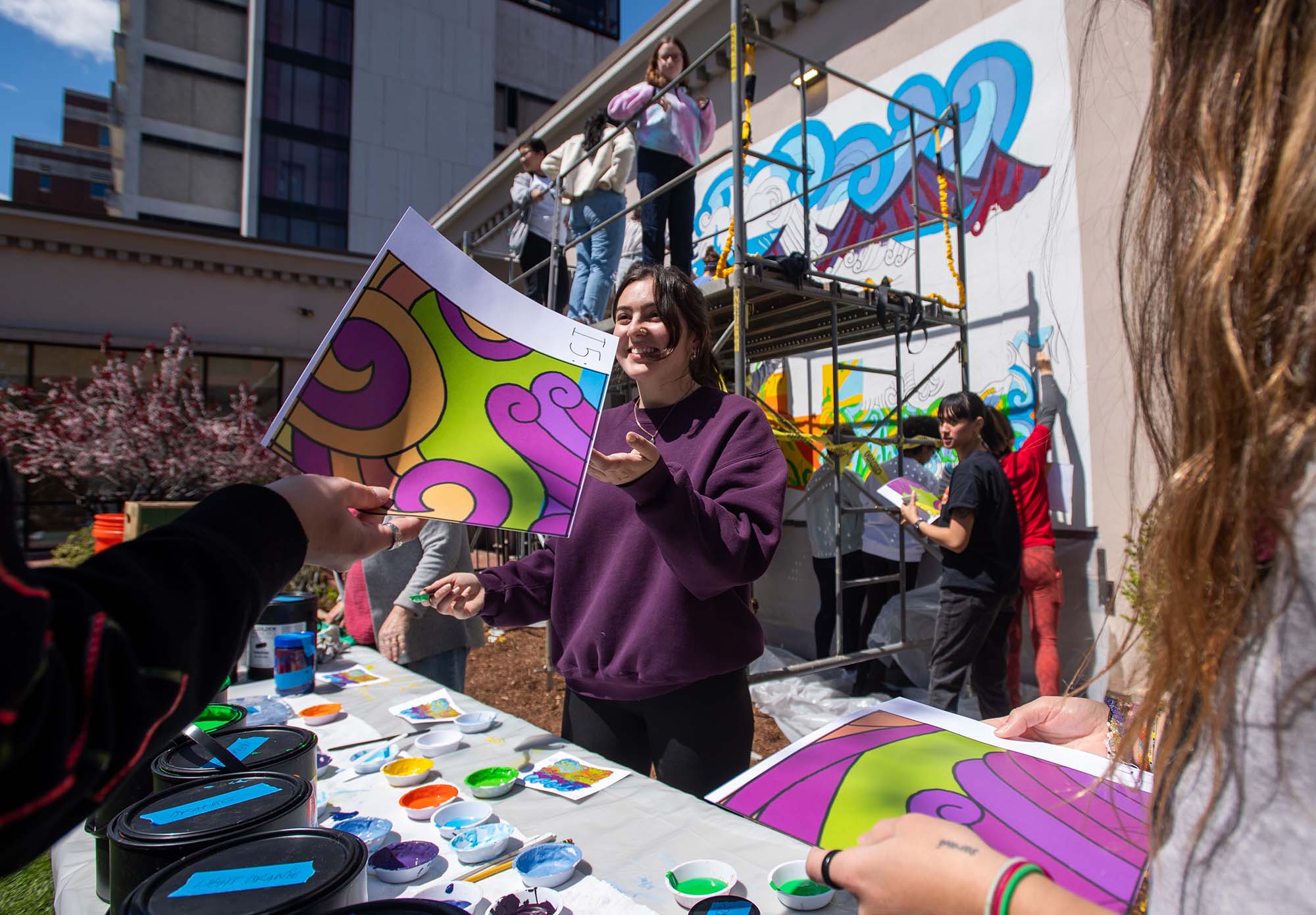 Photo of Artist Halle Cooper, a young white woman with brown hair and wearing a purple sweatshirt, at the painting of her collaborative mural on the side of 565 Commonwealth Ave. She talks to people holding colorful printouts of sections of the mural and stands in front of a table filled with various pots of paint colors.