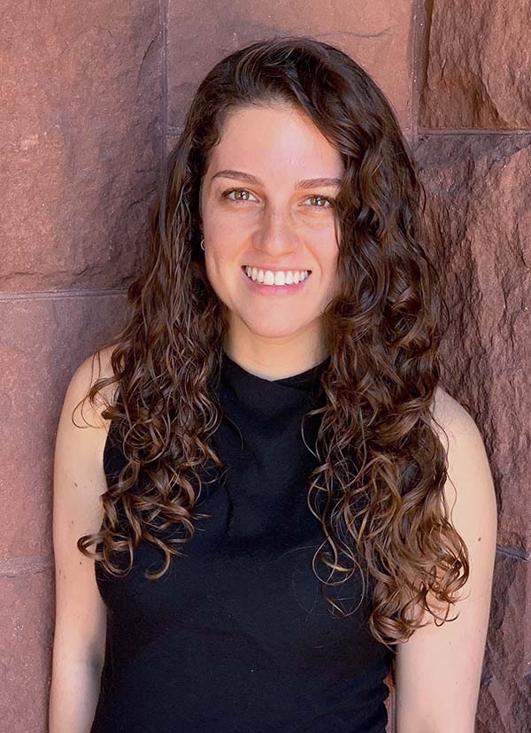 Photo of Neda Khoshkhoo, a light-skinned woman with long brown curly hair. She wears a black sleeveless shirt and smiles as she poses in front of a brick-colored wall.