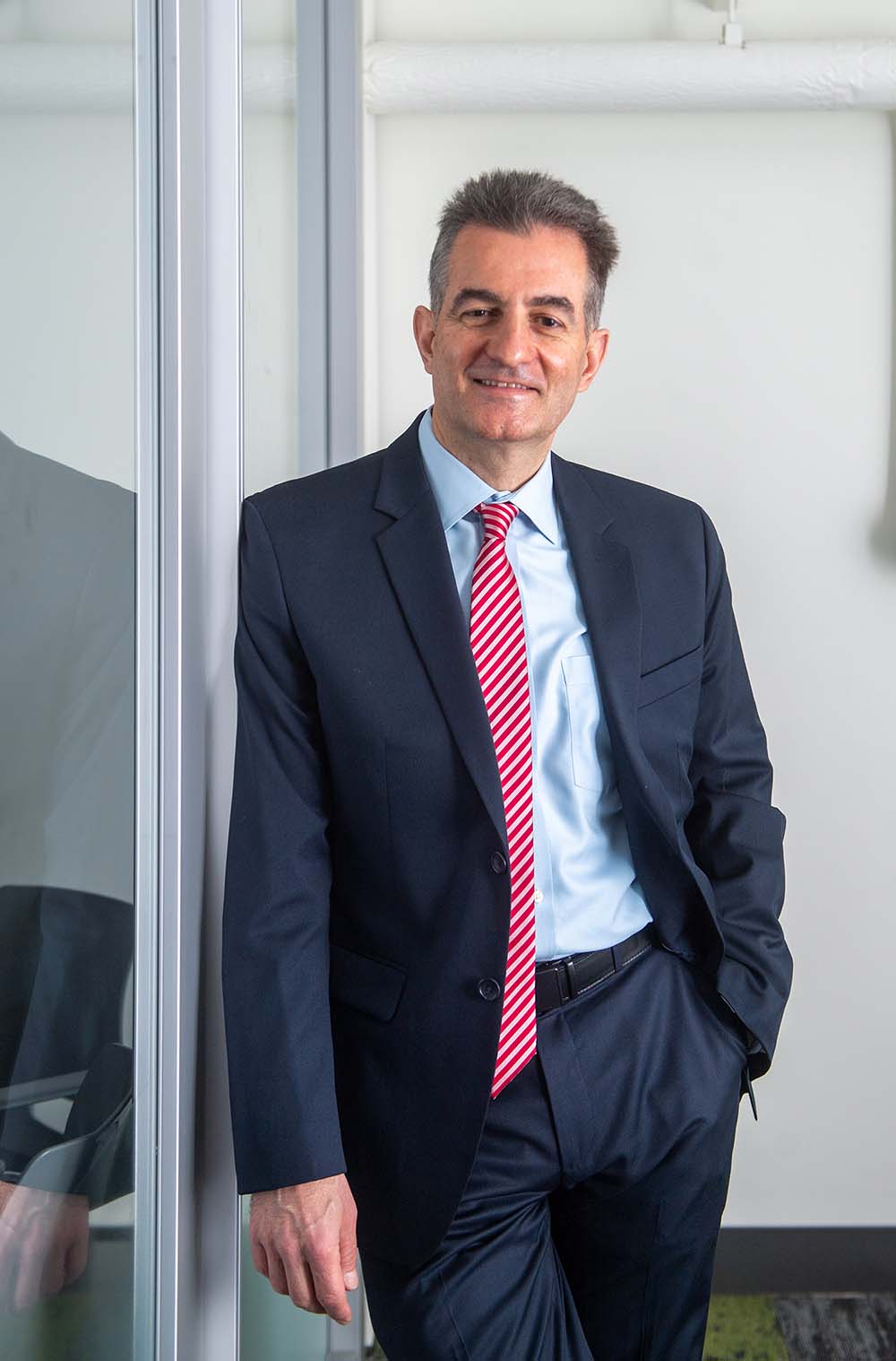 Photo of ENG professor Ioannis Paschalidis, leaning against a glass door and slightly smiling. He wears a dark blue suit, light blue shirt, and red and white striped tie. He is a light-skinned middle-aged man with short brown and gray hair.