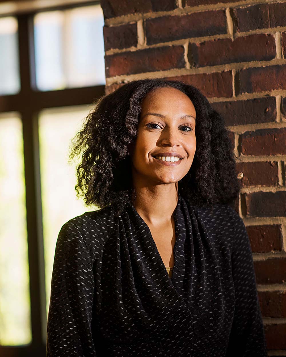 Photo of Clinical Assistant Professor in Health Science, Hagere Yilma, posing for a photo. A Black woman with shoulder-length black hair styled in a twist-out hairstyle wearing a black, tunic-style blouse stands in front of a brick wall and smiles to the camera.