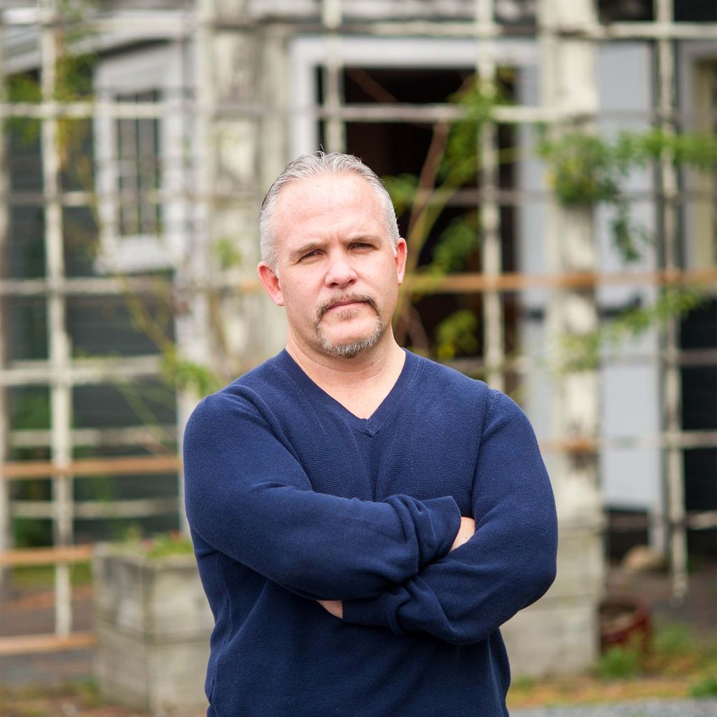 Casey Sherman, a white man with a goatee stands with arms crossed in a blue sweater