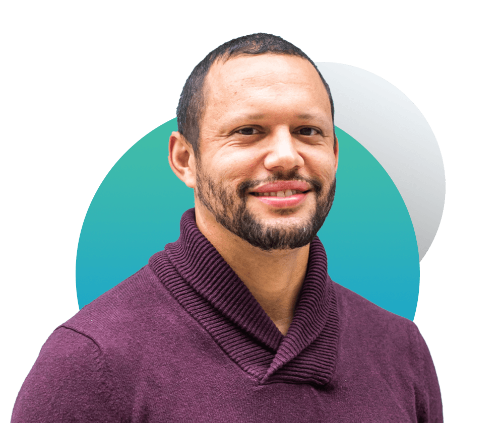 Portrait of Douglas Densmore, a youngish African American man who slightly smiles and wears a crimson-colored quarter zip. He has short brown hair. He is framed by two decorative gradient circles.