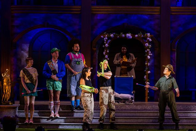 Photo of the company of Much Ado About Nothing during a dress rehersal. On stage, seven people people are seven. At right, a person dressed as a park ranger looks upset and shakes a baton at young people dresses in camoflauge pants and green vests at right. Behind those folks, what appear to be cub scouts are seen. At the back, underneath a trellis, a Black woman reads from a binder.