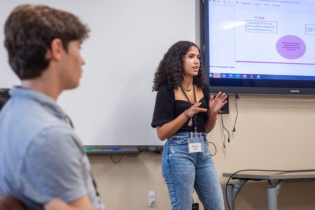 Photo of High schooler Hanna Gialil presenting at a Shark Tank style pitch competition as part of the PopHealth 2 week summer program July 15 at MED. Gialil is a young woman of color with shoulder-length, curly dark hair and light brown skin. She wears hi-rise jeans, a black blouse, and a white name lanyard. Behind her, a screen is seen with a power point presentation; the slide is cut off. A young White man in a light blue polo is seen blurred in the foreground.