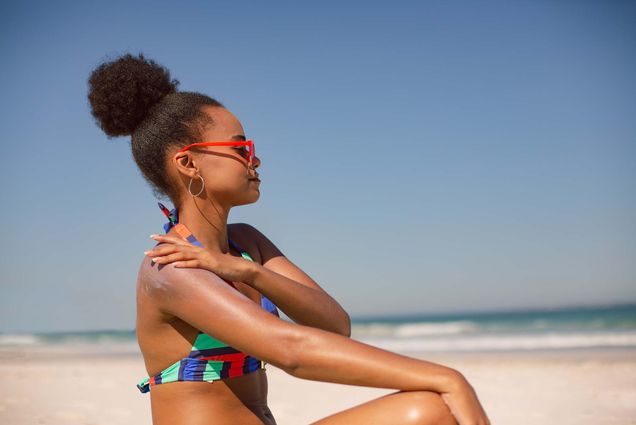 Photo taken from the side of a Black woman in a colorful, striped bikini applying sunscreen lotion on her right shoulder at beach in the sunshine. She sits on the ground and looks towards the water as she applies sun screen. She wears red sunglasses and silver hoop earrings. A clear blue sky and small waves crashing can be seen blurred in the background.