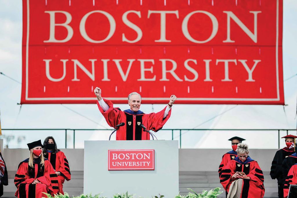 Photo of JR Hipple extending his arms outwards at the podium at graduation; he wears the ceremonial red robe and has a large Boston University sign behind him.