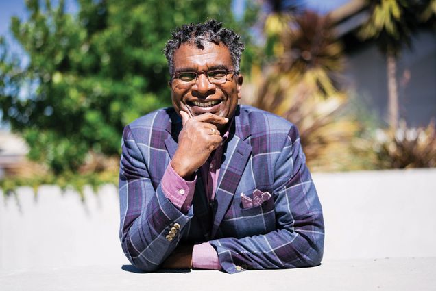 Portrait of Anthony Harrison who wears a blue and purple plaid blazer and rests his chin on his hand and smiles.