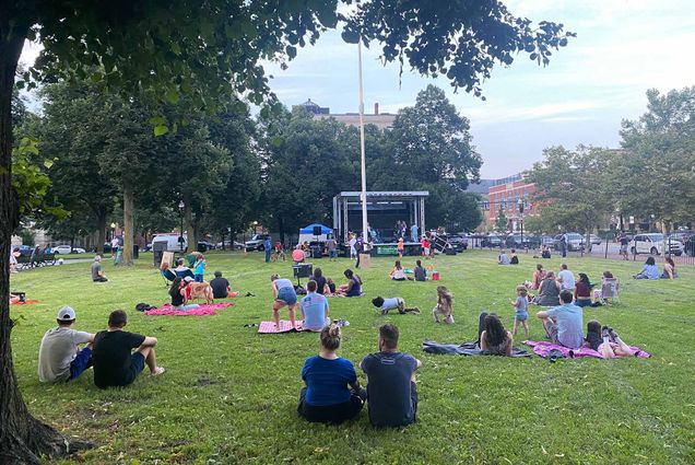 Small groups of people sit on a grassy lawn in Boston in front of an outdoor bandshell