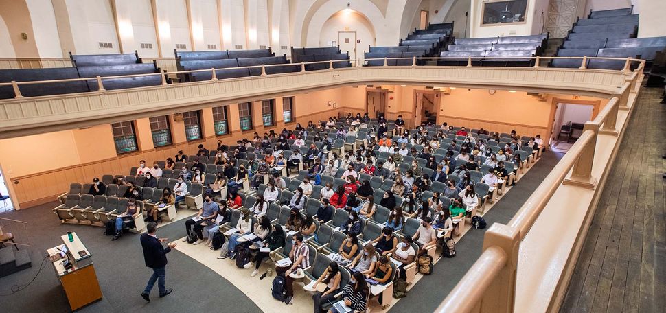 Wide photo of Uwe Beffert’s cell biology class September 2, 2021 in Morse Auditorium, when masking, but not social distancing, was required. The auditorium is two levels: the bottom floor has gray-blue chairs and carpeting, and the second level has black, wooden benches. The space is airy and light, and the bottom is filled with students holding notes and laptops on their laps as they look towards the front, where the lecturer, in a black jacket, is standing in front of a podium.