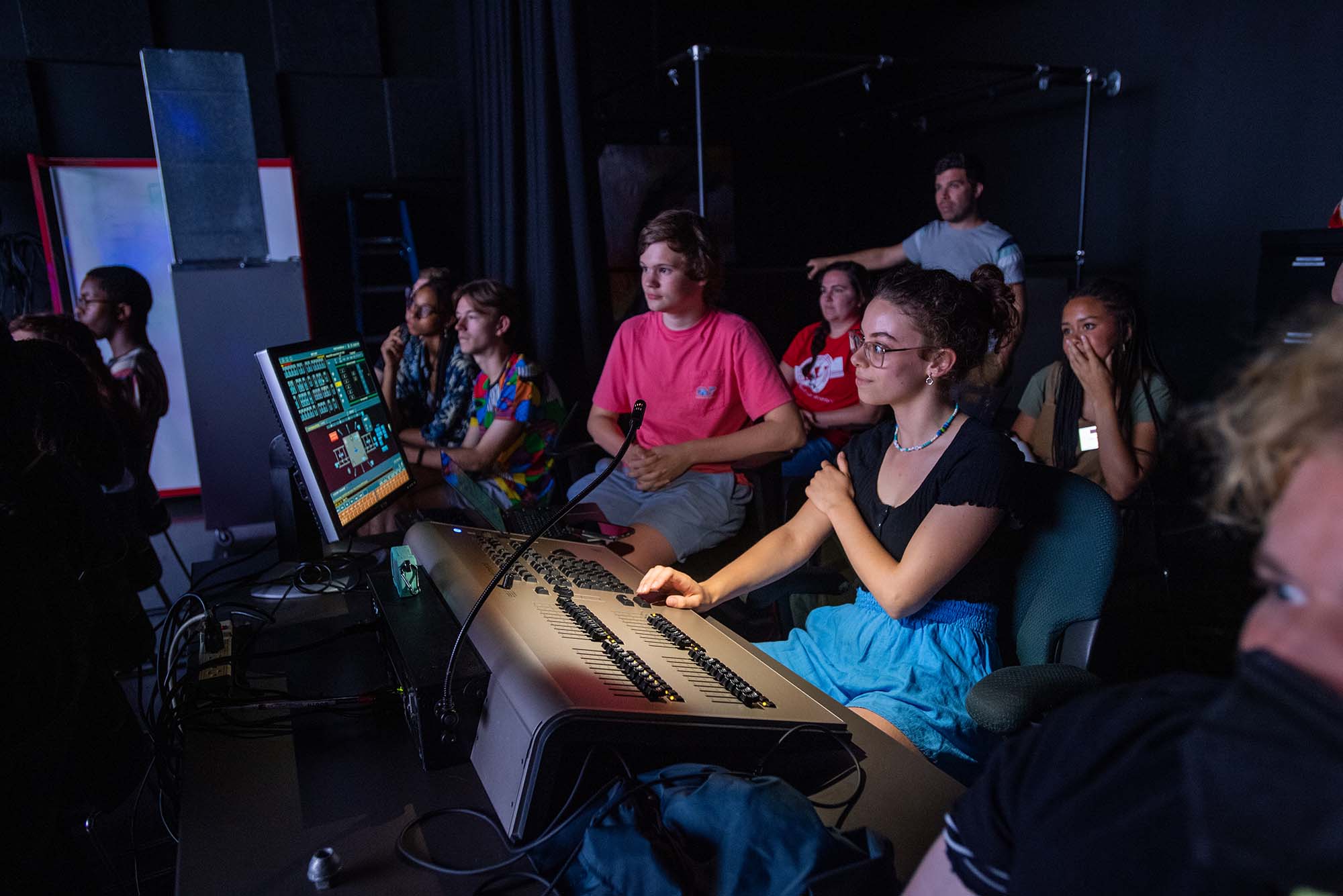 Photo of Nora Kempner (center) a 17-year-old high school student from Medway, Mass., who is enrolled in the BU Summer Theatre Institute’s design program. Kempner is a young White woman with wavy hair, pulled back, wearing a black t-shirt and blue shorts. She sits at a table with lighting controls and a screen and smiles as she shows her lighting scene design with classmates. Other students can be seen seated behind her in the darkened room.