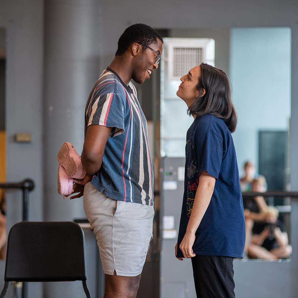 Photo of Joshua Asuru (left), a 17-year-old high school student from Alabama, and Sofia Olona, a 17-year-old from California, perform a scene during a BUSTI acting class. Asuru is a young Black man with short hair, he wears glasses and blue, yellow and gray stripped shirt, and gray shorts. He has a joking facially expression and holds two pink crocs behind his back. Olona has short brown hair and wears a long blue shirt and black bottoms. Asuru and Olona stand face to face, both smiling.