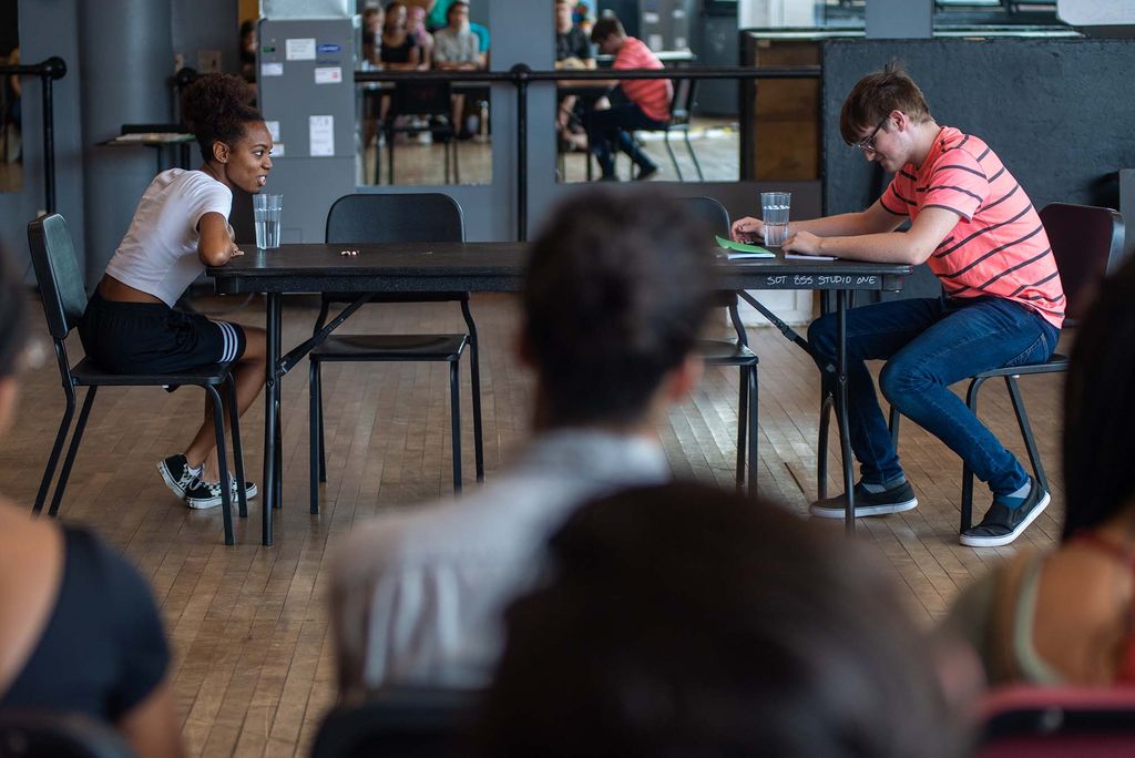 Photo of BUSTI students Ariel Phillips (left) and Colin McCabe seated at a black table in black chairs performing a scene during an acting class. Phillips is a young Black woman with her hair pulled back, who wears a white t-shirt, black shorts and sneakers. McCabe wears a pink shirt with black stripes and jeans. Other students can been seen, blurred, seated in the foreground and reflected in mirrors at the back of the room.