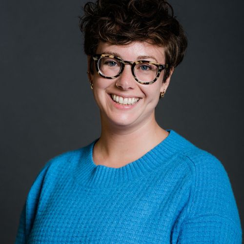 Photo: Headshot of Molly Callahan. A white woman with short, curly brown hair, wearing glasses and a blue sweater, smiles and poses in front of a dark grey backdrop.