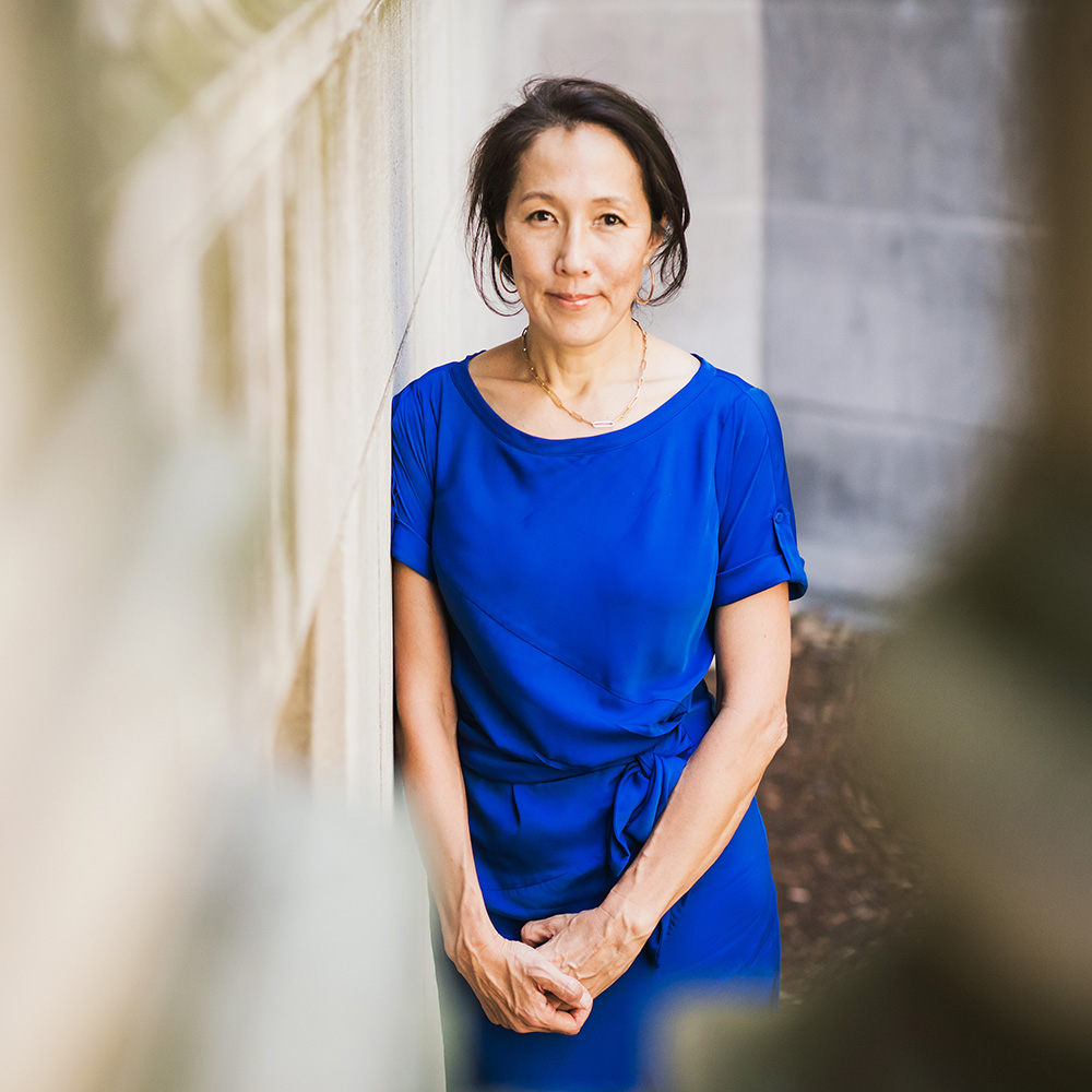 Photo of Christina Lee, a SSW associate professor and member of the addiction faculty at BU’s Grayken Center at BMC. A woman with black hair tied back into a small bun leans against a pale yellow wall and looks at the camera as she poses for the photo. She wears a blue blouse.