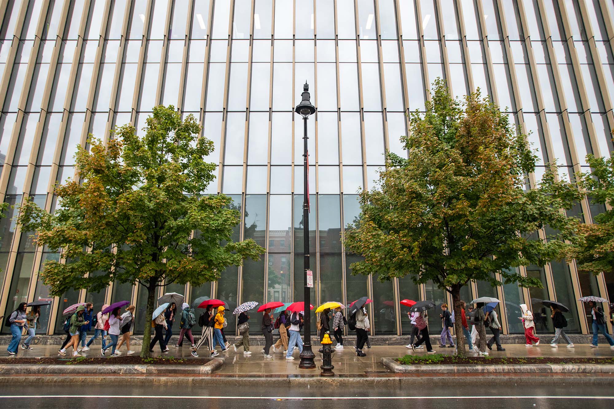 Umbrellas add a touch of color to a dreary Thursday, as Terriers wend their way past the Kilachand Center for Life Sciences & Engineering. Rain dumped nearly an inch on 波士顿, and Friday is expected to be much cooler, with a high of only 60. Could be fall has arrived. September 22 photo by Cydney斯科特. Students walk in front of a large modern building with umbrellas in rainy weather