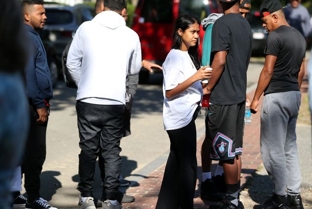 Photo: A group of migrants huddle on a sidewalk in front of St. Andrews Episcopal Church. A young Latina woman wearing a large white shirt holds a white cup and looks off as other Latinx individuals stand and look off around ehr.