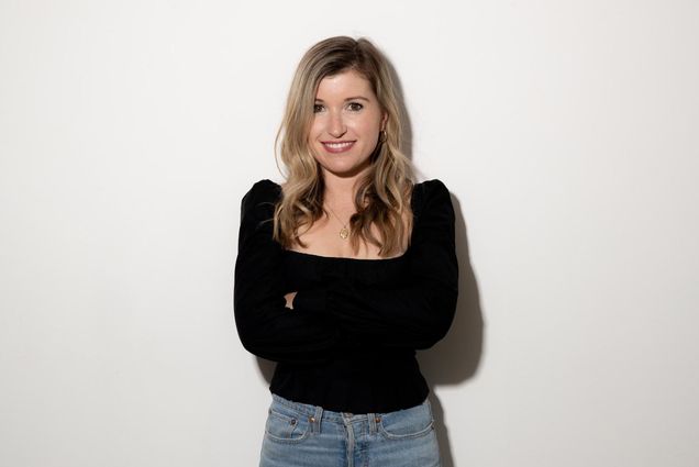 Photo: Studio headshot of Sarah Moret. A young white woman wearing a long-sleeved black shirt and jeans smiles in front of a white studio backdrop.
