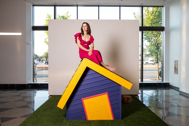 Lowell-based artist Jaina Cipriano creates installations that evoke the joy and pain of childhood nostalgia. Here she sits atop one of her creations, a tilted blue house with a yellow roofline and door. She is wear a red dress.