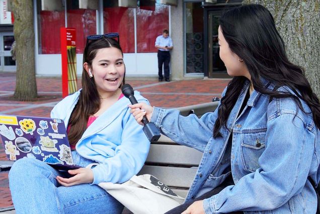 Photo of Irene Kim interviewing a student in front of the BU student union. Two female students sit on a bench facing each other. The student on the right holds a microphone up as the student on the left speaks into it.