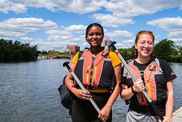Photo of Isabelle Yap (Sargent'23) , (left), and Genesis Veiassa (Sargent'24) standing on a dock along the Charles River. The Harvard Bridge is seen behind them in the distance; the water and sky are blue with puffy clouds. Yap is a young Black woman with her dark hair pulled back. She has a backpack slung on one shoulder and wears a yellow top and black spandex. Veiassa is a white Woman with long red hair pulled back into french braids, she wears a black t-shirt and gray shorts. Both smile, wear orange, black and gray life jackets, and hold canoeing paddles across their chests.
