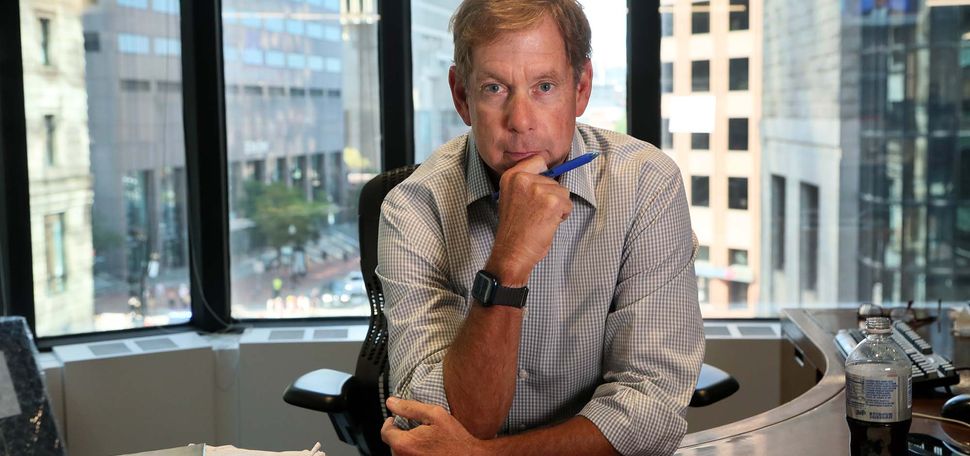 Photo of Brian McGrory, in his office at the Boston Globe. McGrory is a middle-aged White man with short blondish hair. He poses with his hand on his chin and holds a blue pen. He has a large desk that curves around to the back and his office has a large window that looks out onto downtown Boston. Papers and diet pepsi are seen in the foreground.