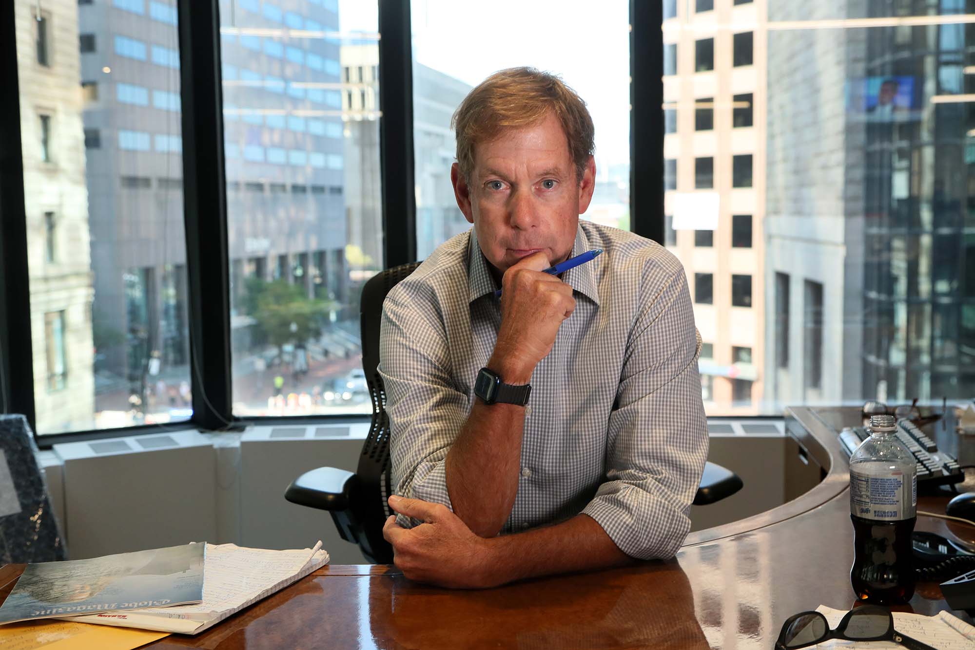 Photo of Brian McGrory, in his office at the Boston Globe. McGrory is a middle-aged White man with short blondish hair. He poses with his hand on his chin and holds a blue pen. He has a large desk that curves around to the back and his office has a large window that looks out onto downtown Boston. Papers and diet pepsi are seen in the foreground.