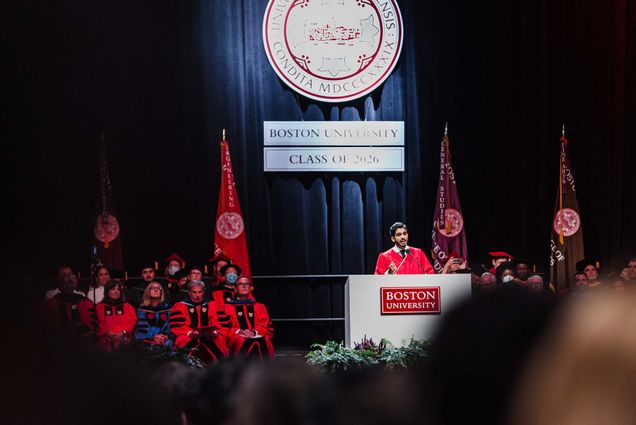 Photo of Dhruv Kapadia giving a speech at BU's 2022 Matriculation. A young South Asian man wearing red robes stands at a podium in front of a stage decorated in black curtains and with a large BU insignia hanging behind him. Blurry students are seen in the crowd in front of him, as other robed officials sit on stage to his left.