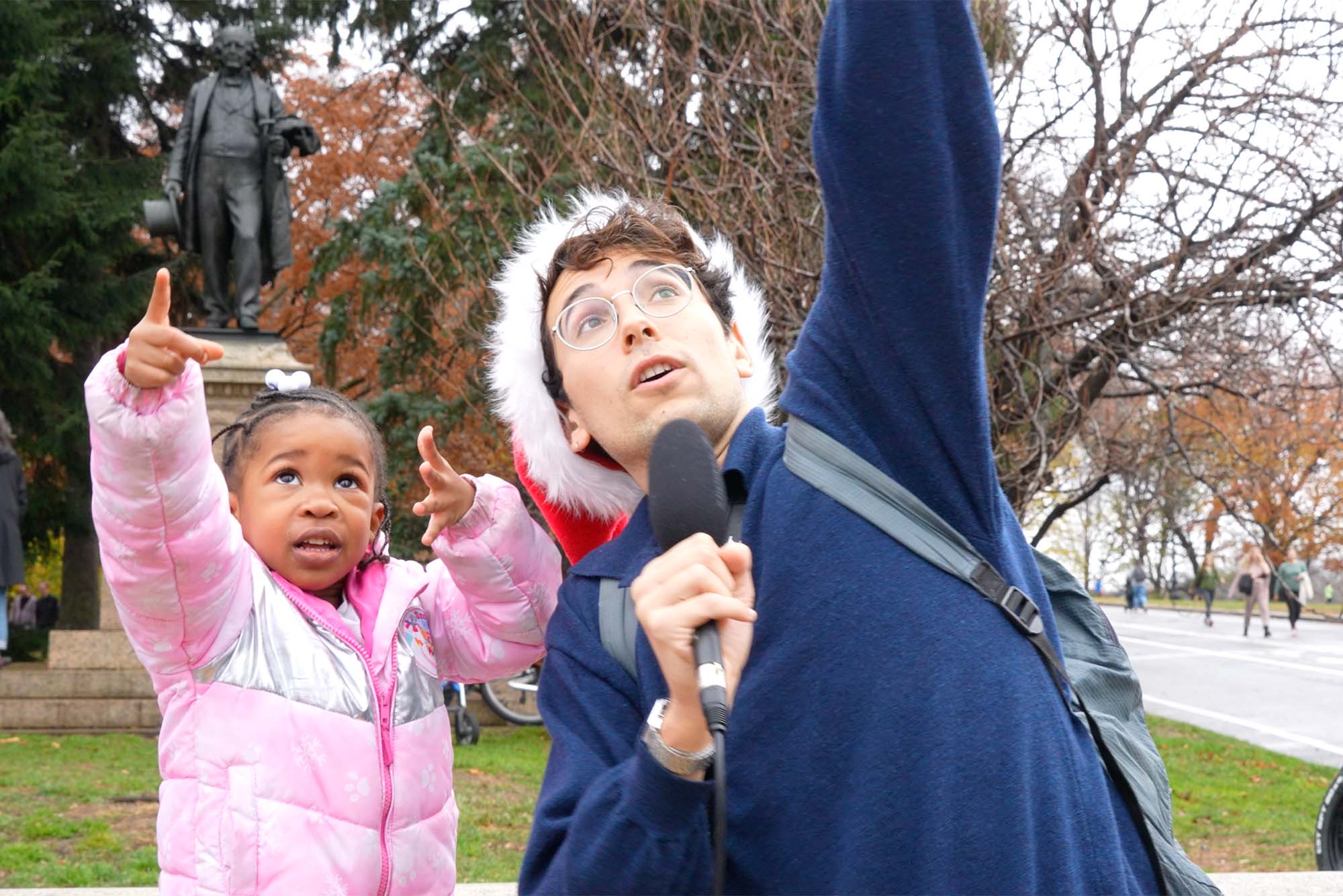 Image: screenshot of Julian Shapiro-Barnum, a young white man wearing glasses, a navy sweater, and a red Santa hat, interviewing a small child Julian holds a black microphone to his face as he and the child point to the sky.