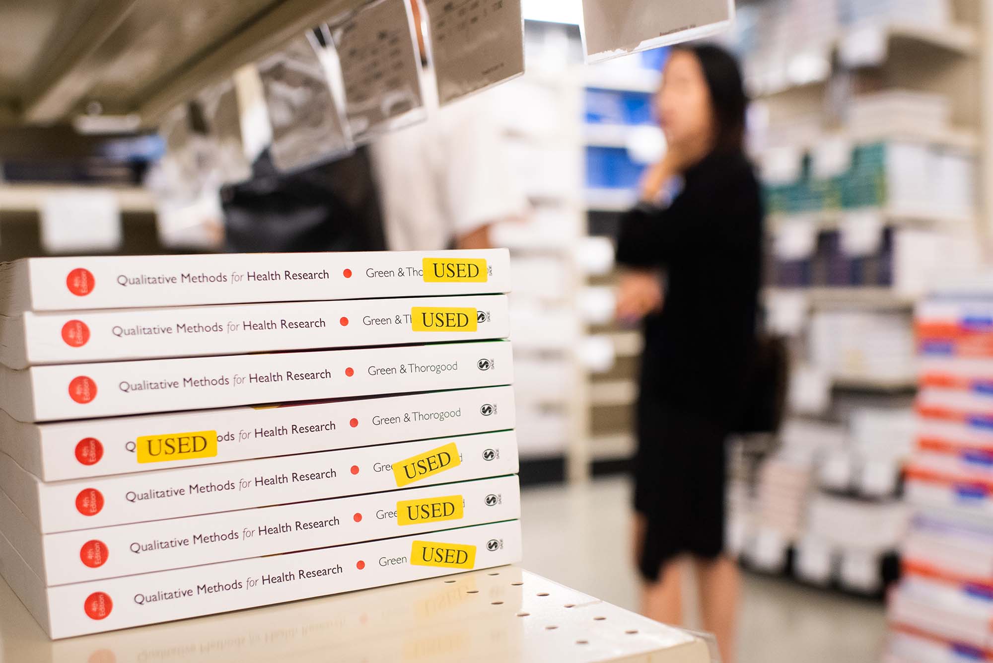 Photo of a stack of books with a yellow "USED" sticker on them. The stack of white books are "Qualitative Methods for Health Research" from "Green & Thorogood." The books sit on a tan industrial shelf and are in focus. Someone in a black dress stands in aisle of the bookstore, blurred, in the background.
