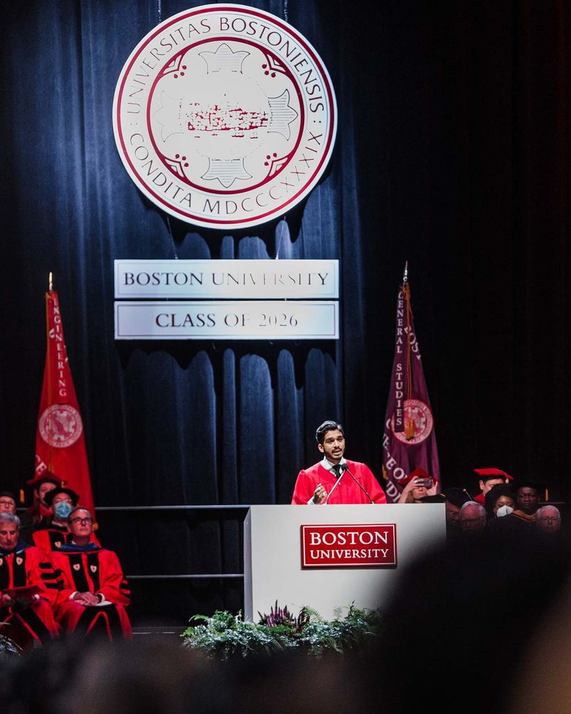 Photo of student speaker Jason Campbell-Foster speaking at a gray podium with a red Boston University sign during matriculation. He wears a red and black ceremonial robe and the white and red BU seal is seen behind him. He is on stage with faculty, and the backs of students' head are seen blurred in the foreground.