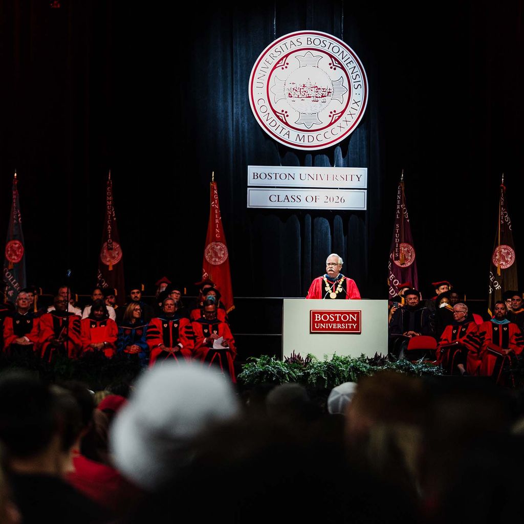 Photo of president Brown speaking at a gray podium with a red Boston University sign during matriculation. He wears a red and black ceremonial robe and the white and red BU seal is seen behind him. He is on stage with other faculty, and the backs of students' head are seen blurred in the foreground.