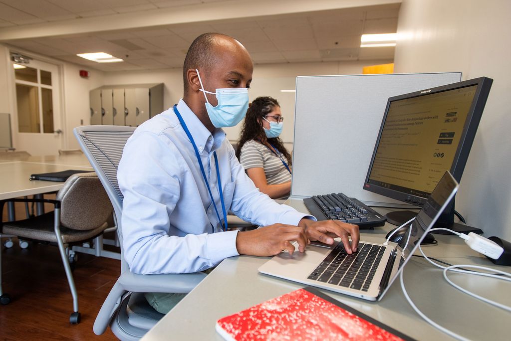 Photo of pulmonary fellow Nathan Mesfin, with fellow fellow Mallory Lesieur at MED September 1. They both wear blue face masks and sit at computers.