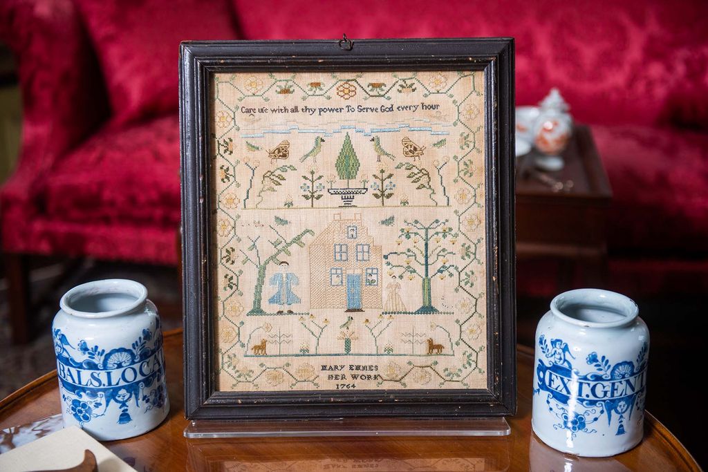 Photo: A framed, old needlework is displayed in a black frame. Two small white and blue vases sit on either side of it on a wooden table.